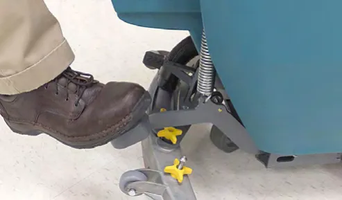 A foot pressing the pedal to lower the scrub head, beside the yellow adjustment knobs