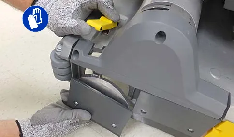 Hand unfastening the yellow latch to remove the idler plate; protective gloves symbol