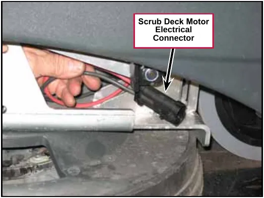 A hand unplugging the black scrub deck motor connector under the machine frame. Label: Scrub Deck Motor Electrical Connector.