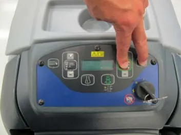 Hand on the membrane switch control panel with two fingers pressing the scrub on/off switch and the vacuum/wand switch, key switch at the lower right