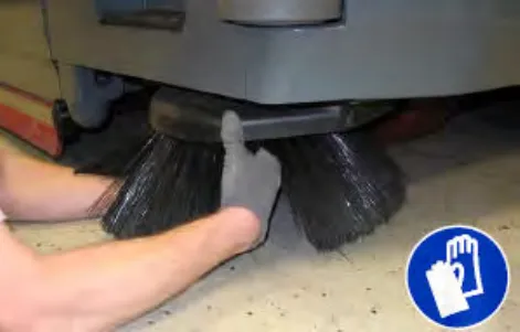 Hand reaching into the center of the sweeping side brush to remove the cotter pin, plus glove symbol