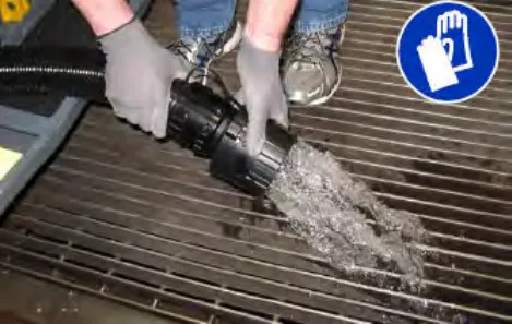 Gloved hands holding the drain hose nozzle over a floor drain as water runs out, with a glove-required symbol at upper right