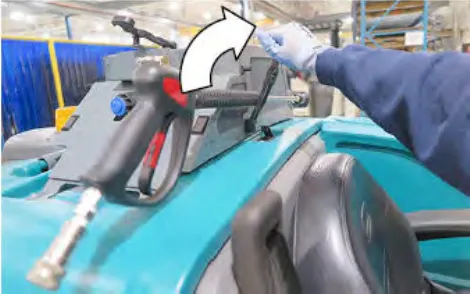 A gloved hand unlatching the tie down on the pressure washer wand, with a curved arrow