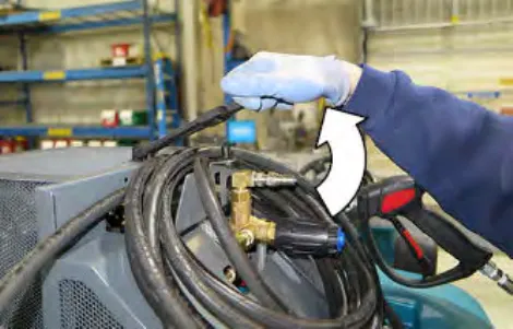 A gloved hand unlatching the tie down on the coiled pressure washer hose, with a curved arrow
