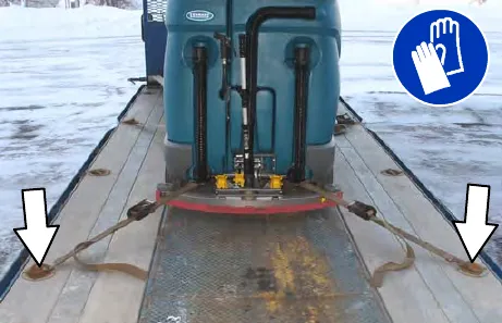 Rear view of the machine strapped to a trailer with two straps, arrows pointing to the trailer tie-down points; gloves must be worn