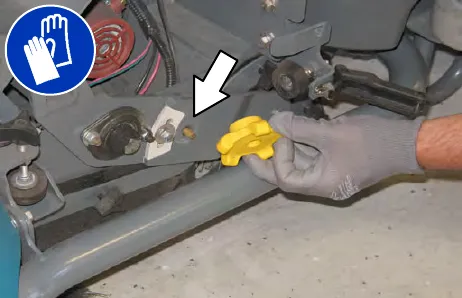 Gloved hand removing the yellow left brush arm knob, with an arrow pointing to its location; gloves must be worn