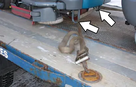 Front of the machine on a trailer, with arrows pointing to the stabilizer legs where the tie-down strap hooks on