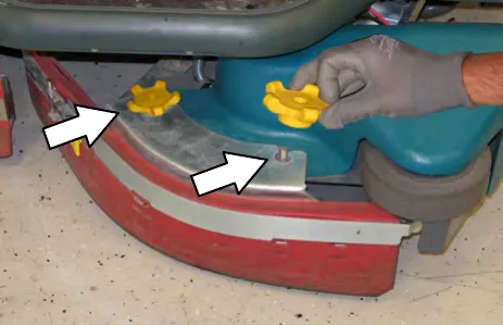 Gloved hand removing the yellow knobs that hold the side brush squeegee assembly, with arrows at the knob locations