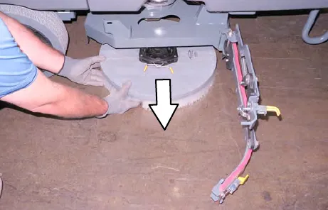 A person pulling the dropped disk brush out from under the scrub head, with an arrow pointing down