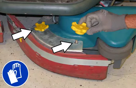 Gloved hand removing the yellow knobs holding the side brush squeegee assembly, with arrows at the knob locations; gloves must be worn